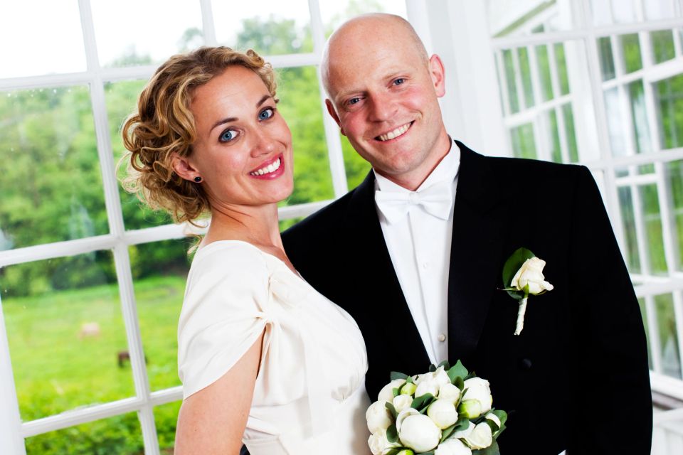 Bryllupsfoto af smilende par med brudebuket, stående elegant foran et vindue med grønne træer i baggrunden.