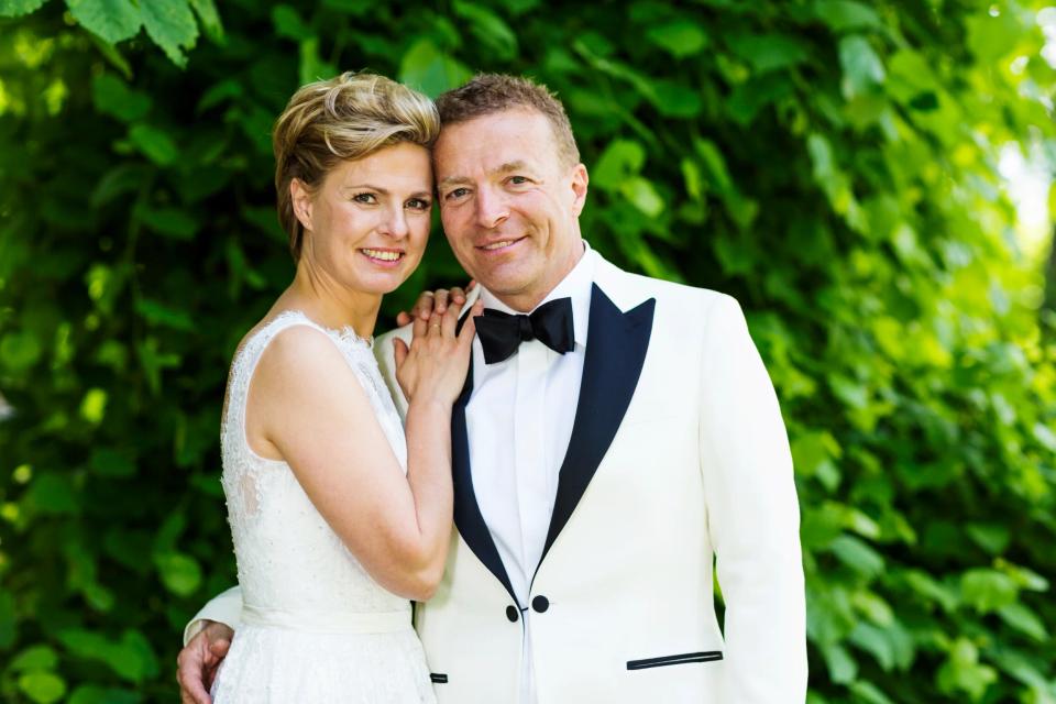 Bryllupsfoto af smilende brudepar tæt omfavnende foran grønne blade i blødt dagslys. Glad stemning.