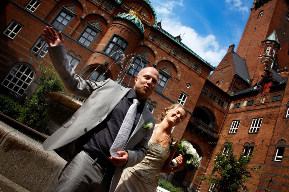 Fejrende brudepar foran Københavns Rådhus. Manden vinker, og kvinden smiler med sin brudebuket. Solskinsdag.