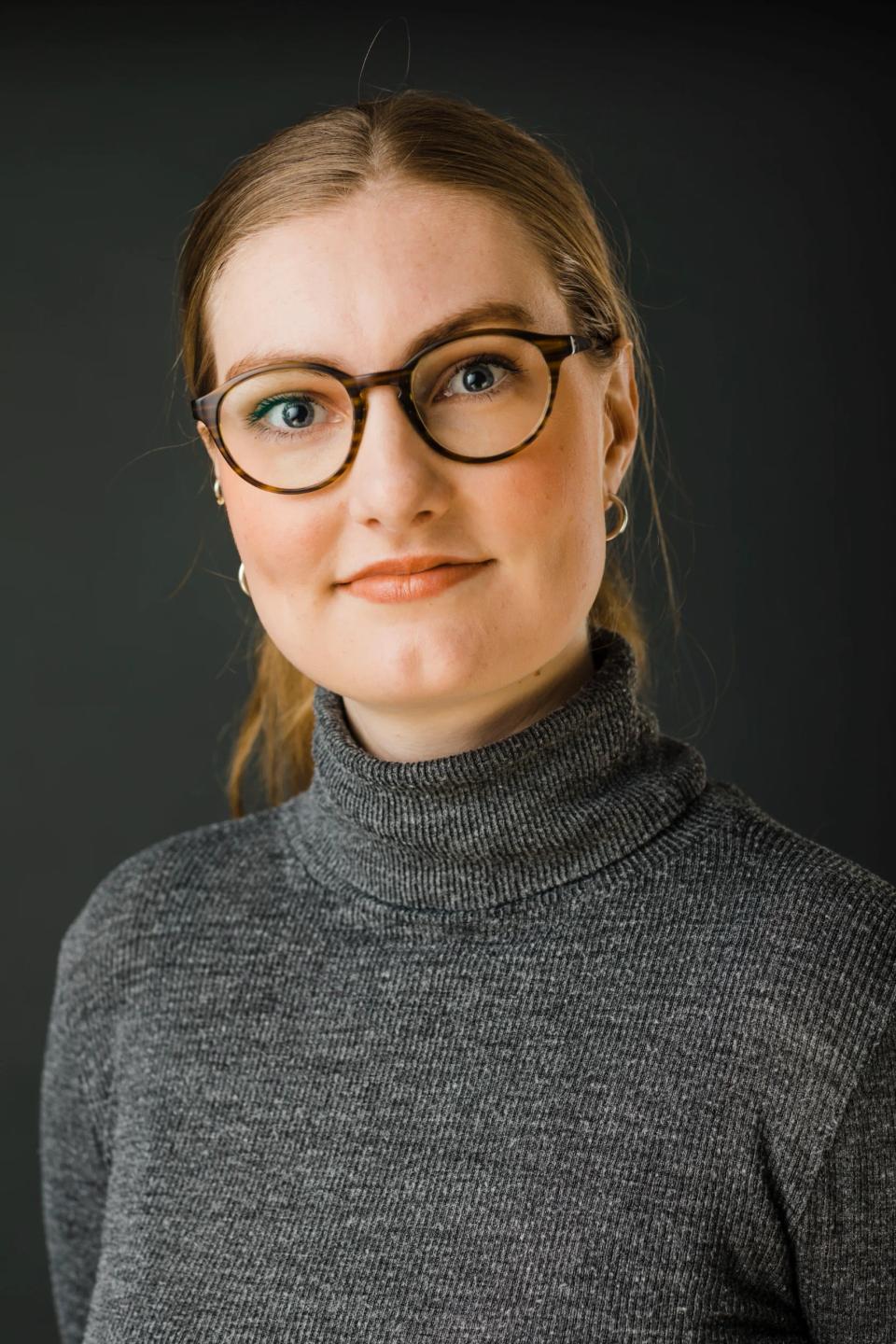 Portrætfoto af smilende kvinde med lysebrunt hår, runde briller og grå rullekrave mod mørk baggrund.