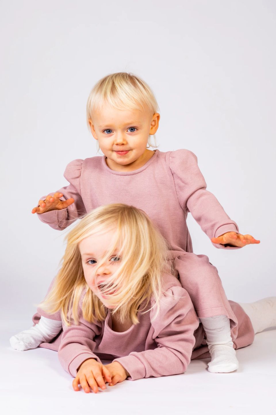 Legesyge børnebilleder: To smilende små piger med lyst hår i lyserøde sæt, den ene kravler på den anden.