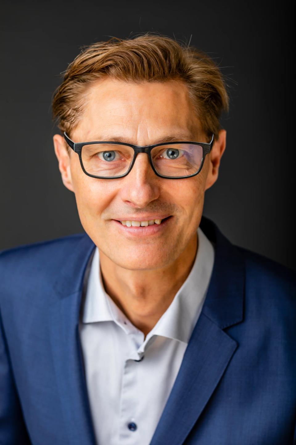 Portrætfoto af smilende mand med briller og blåt jakkesæt i blødt lys, der ser direkte ind i kameraet.
