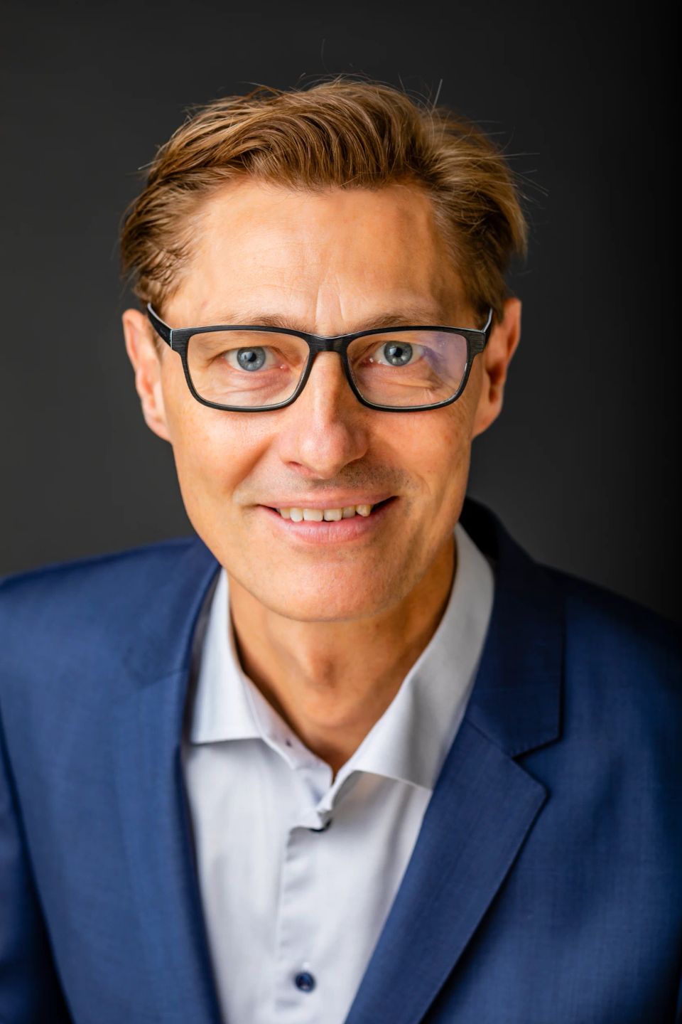 Portrætfoto af smilende mand med briller og blåt jakkesæt i blødt lys, der ser direkte ind i kameraet.