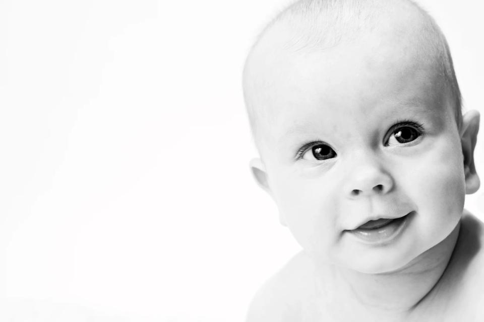 Nærbillede i sort/hvid af en glad baby der smiler nysgerrigt mod beskueren. Et charmerende babyfotografi.