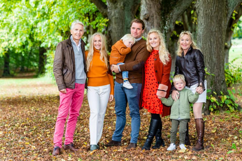 Familiefoto af tre generationer, grøn løvskov med nedfaldne efterårsblade. Glade ansigter i naturligt lys.