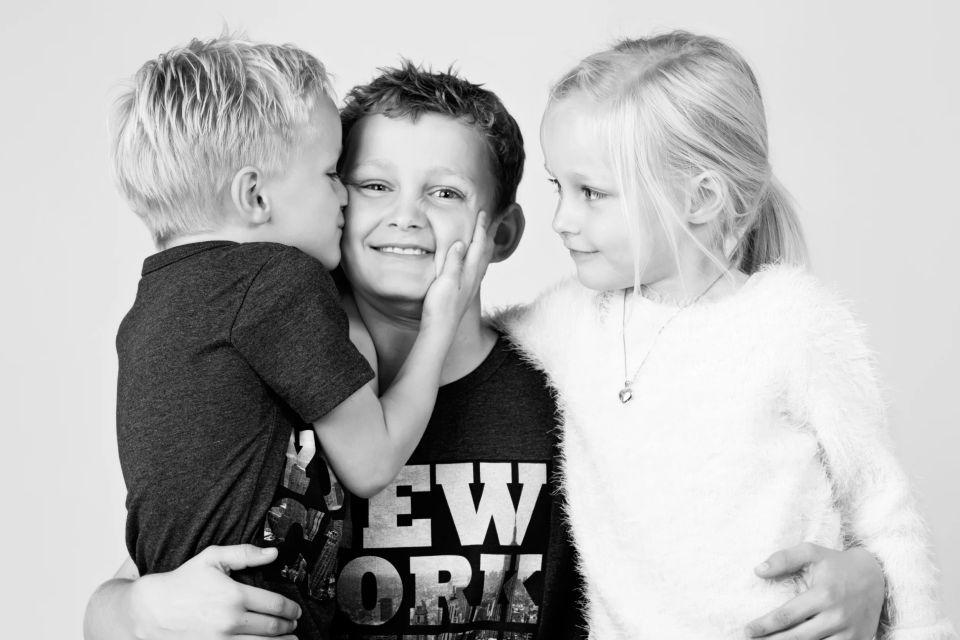 Sort/hvid portrætfoto af tre glade børn, hvor den yngste giver den ældste et kys på kinden. Søskendekærlighed i et børnebi...