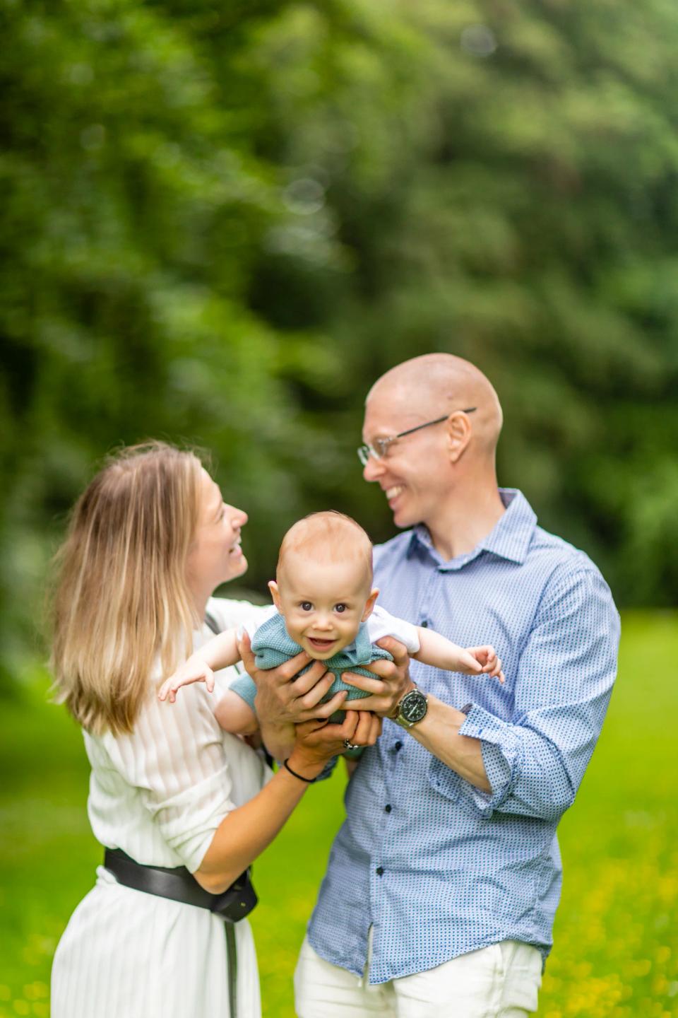 Familiefotografering: Smilende forældre holder glad baby, der strækker armene ud, i grønt sommerlys i Sophienholm.