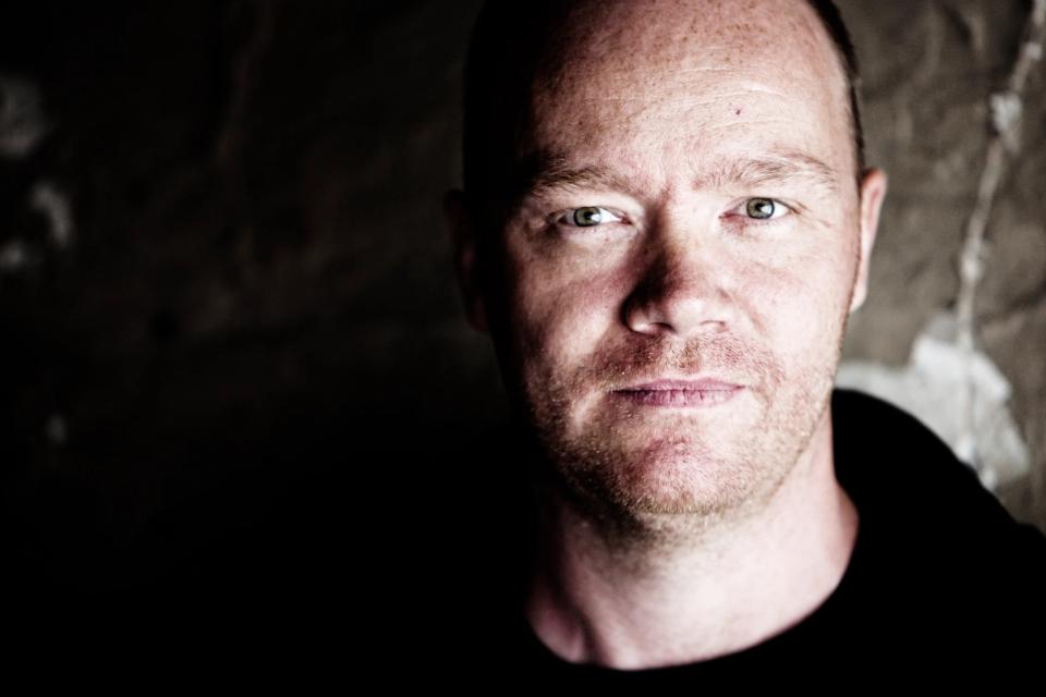 Nært portrætfoto af en mand med skægstubbe, der kigger direkte ind i kameraet, oplyst fra siden i dæmpet belysning.