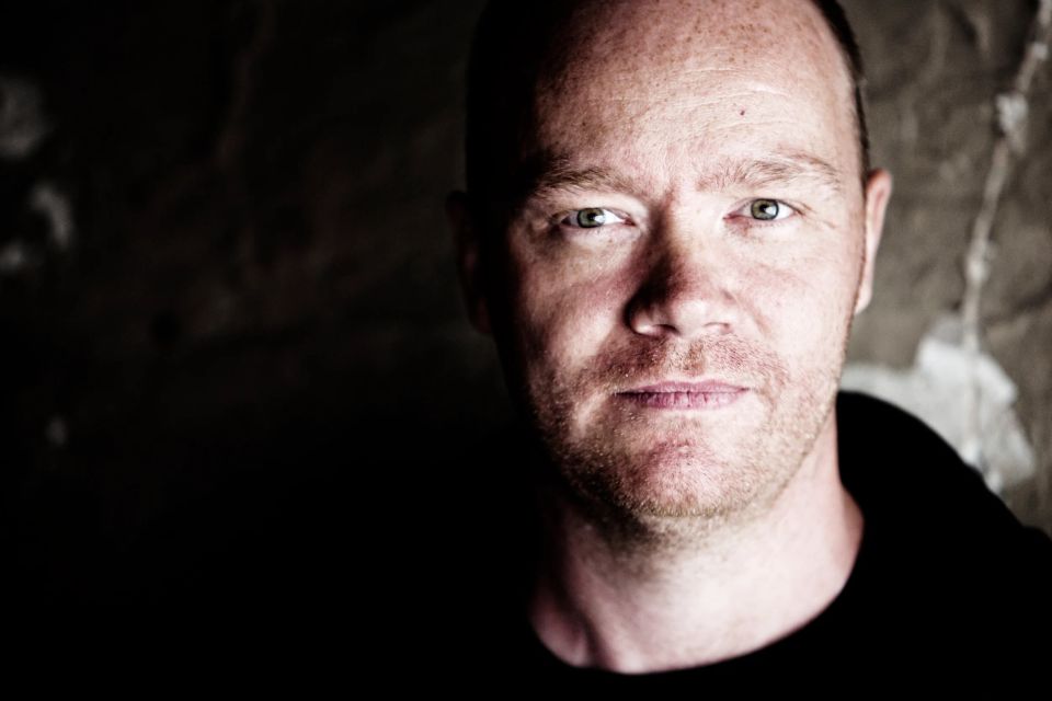 Nært portrætfoto af en mand med skægstubbe, der kigger direkte ind i kameraet, oplyst fra siden i dæmpet belysning.