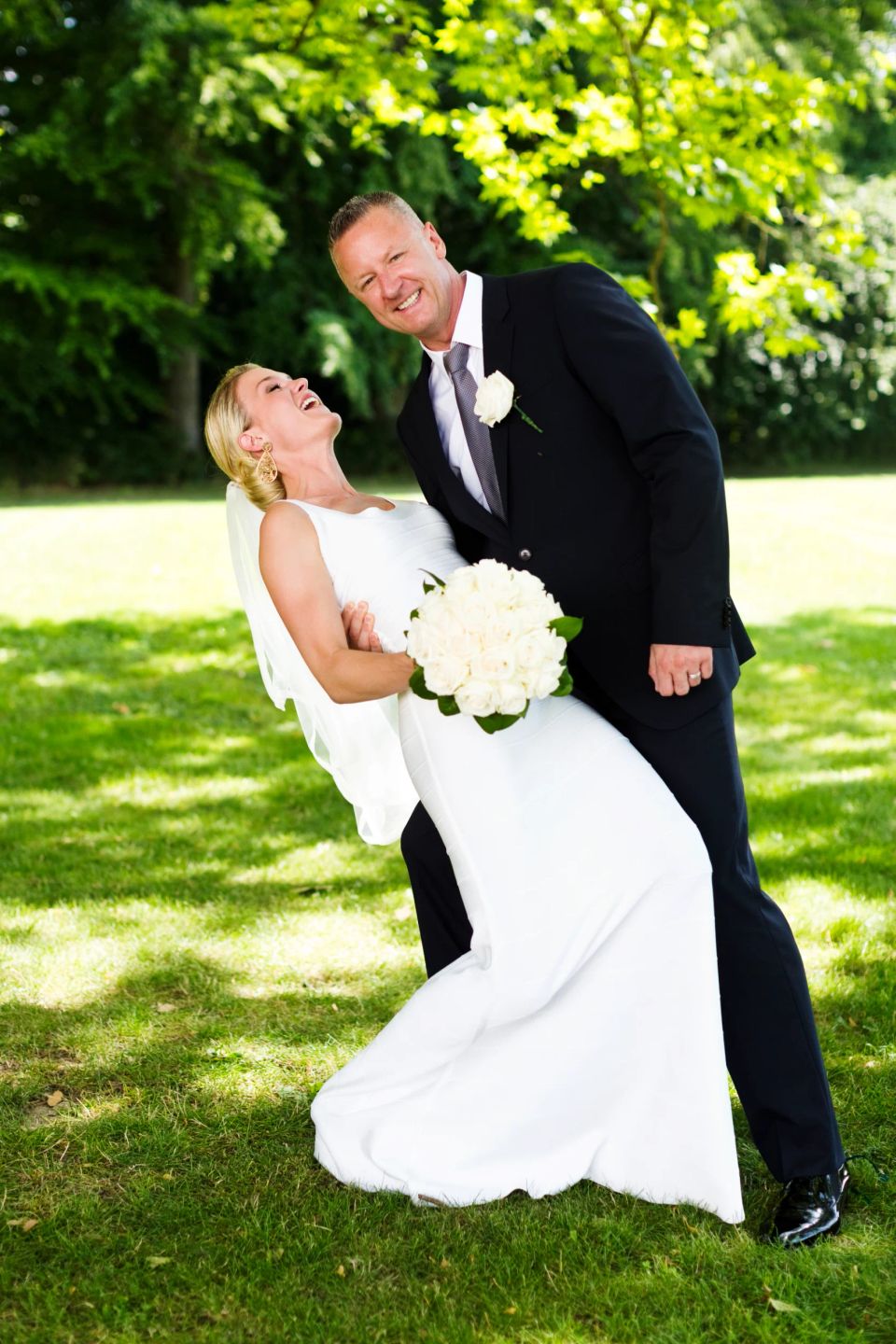 Bryllupsfotografering af grinende brudepar. Bruden læner sig tilbage holdt af brudgommen i grøn natur, solskin og de ser g...