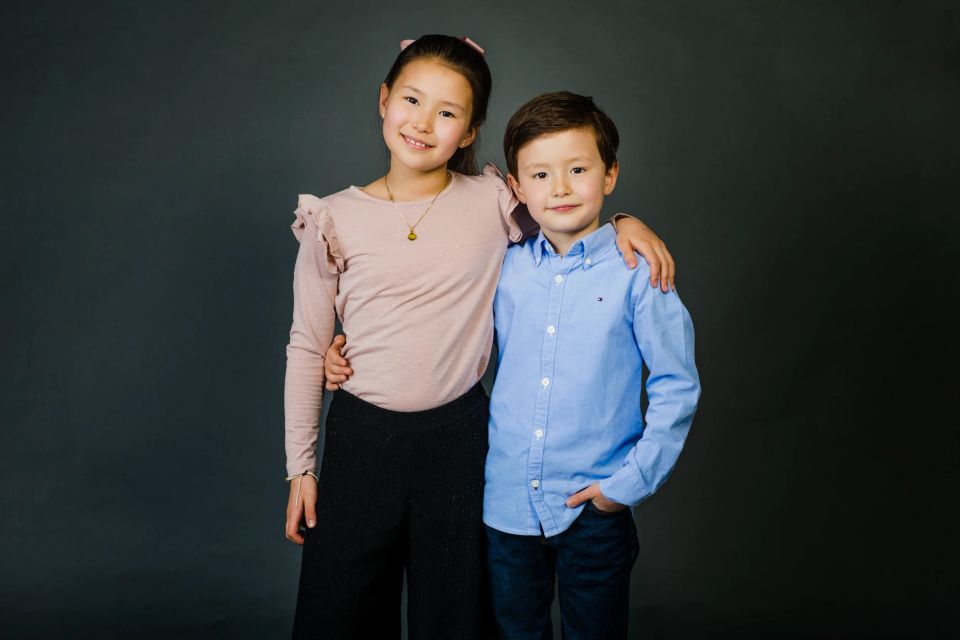 Børn: Søskendeportræt af to smilende børn, en pige og en dreng, poserer tæt sammen foran en mørkegrå baggrund.