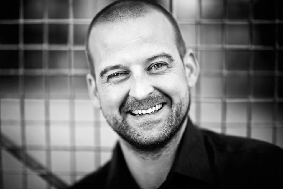 Sort-hvid portræt af skaldet, smilende mand med skægstubbe. Ansigtet er tæt på og baggrunden er sløret.