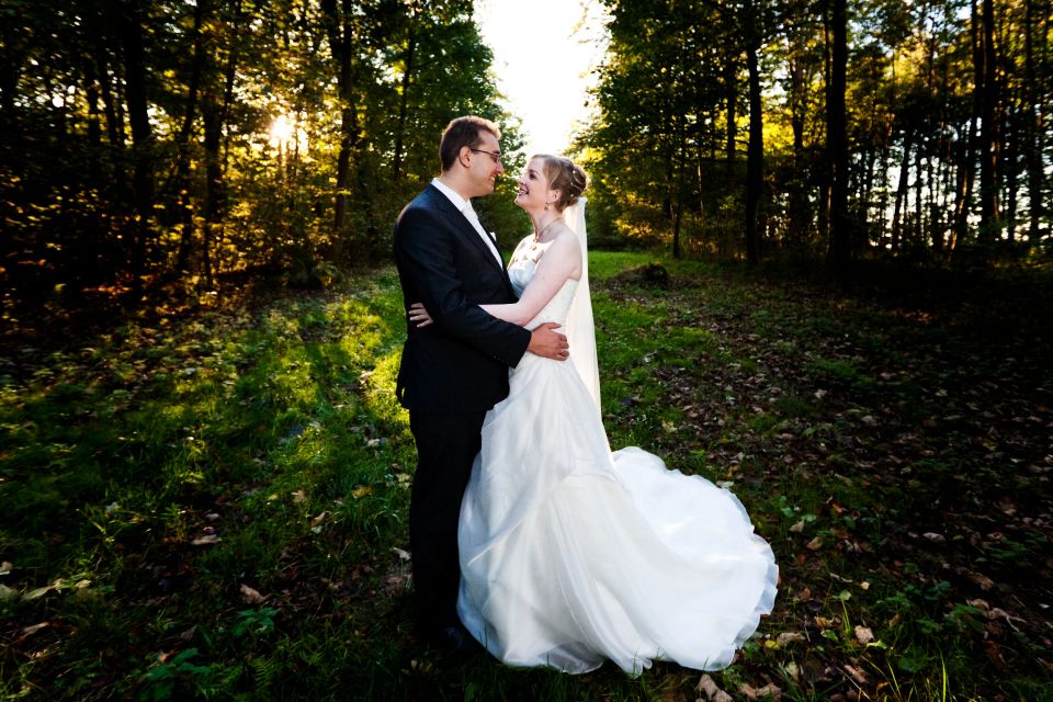 Bryllupsfotografering af smilende par i kærlig omfavnelse i skovbrynet med underskov og solskin i baggrunden.