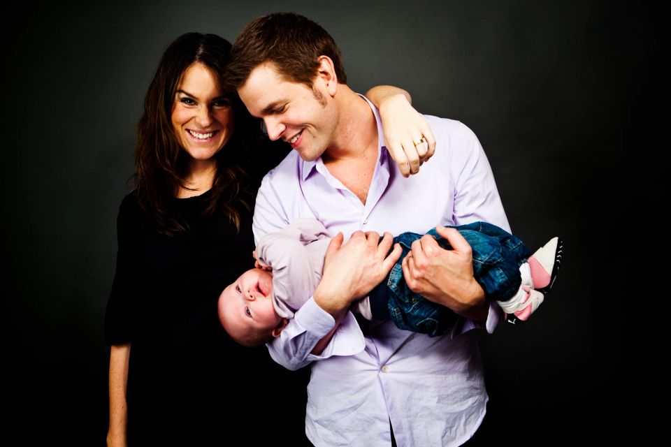 Familiefoto: Forældre smiler varmt, mens far holder baby glædeligt på hovedet mod en mørk baggrund.