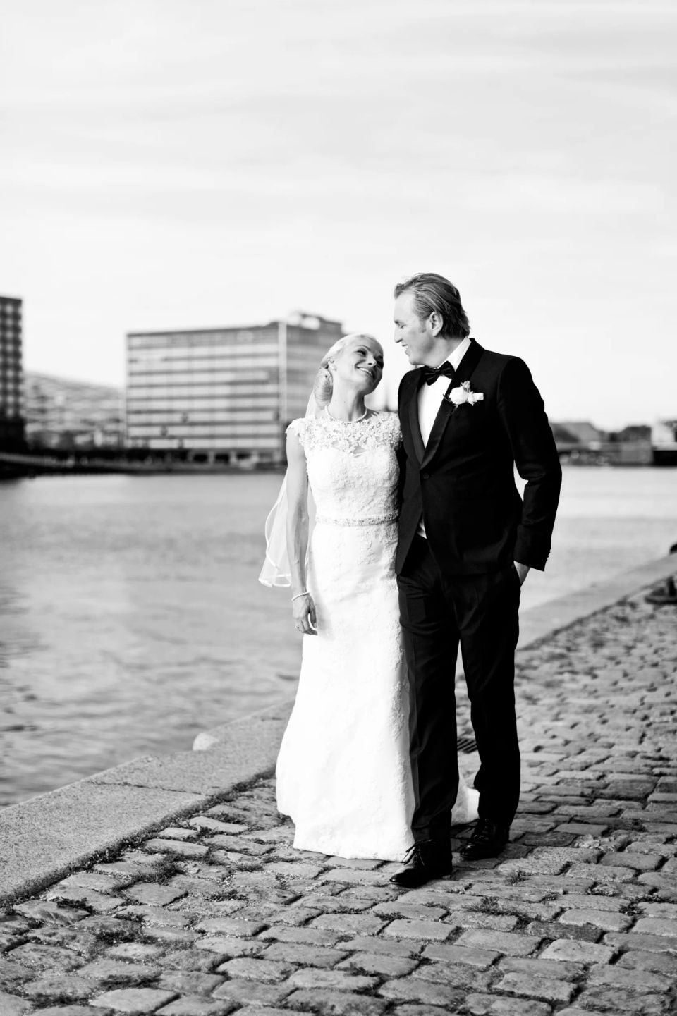Bryllupsfotografering af et smilende brudepar der går langs vandet på brosten, et kærligt blik.