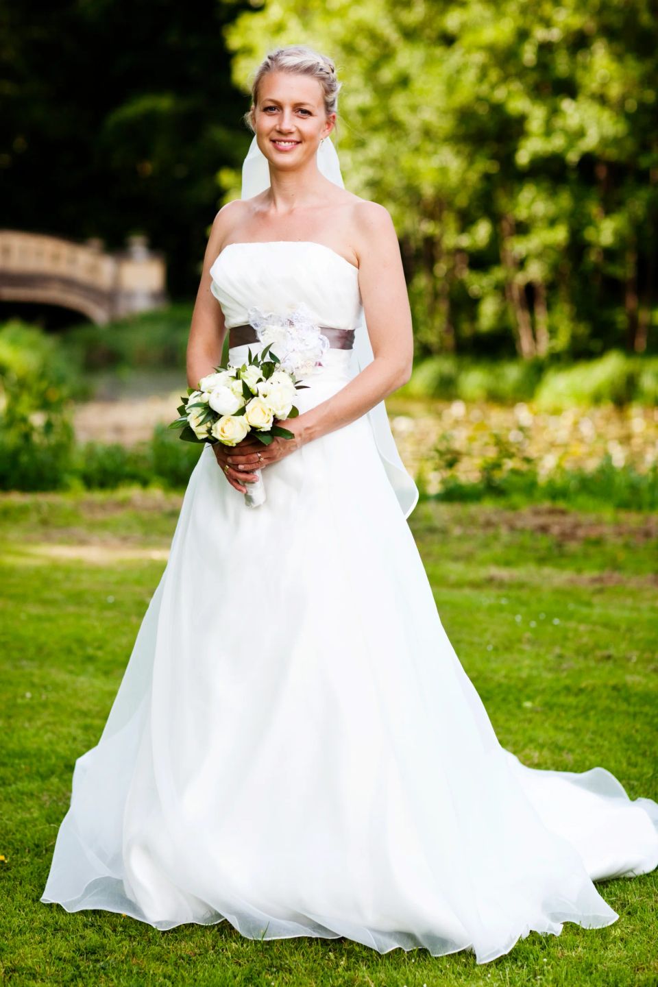 Bryllupsfotografering af glad brud i hvid kjole med brudebuket, stående på græs i Aldershvile slotspark.