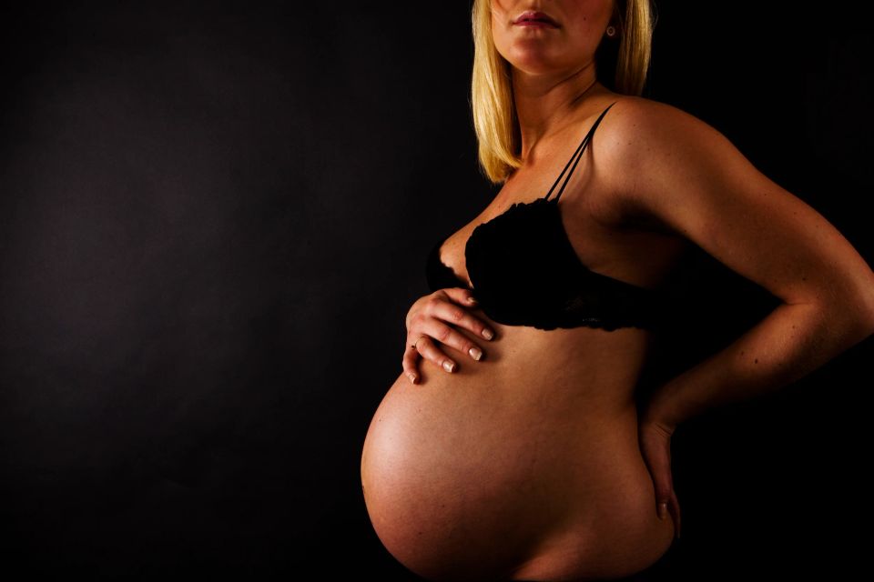 Nærbillede af gravid kvinde i sort blondetop, der holder om sin mave. Dramatisk lys fremhæver silhuetten mod mørk baggrund.