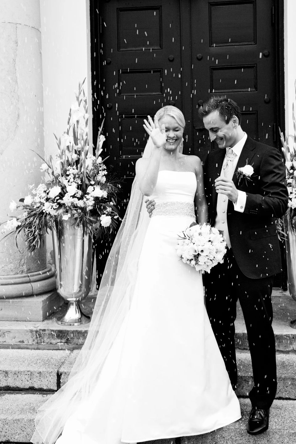 Bryllupsbillede af smilende brudepar efter vielse, omgivet af regn af riskorn. Bruden vinker glad.