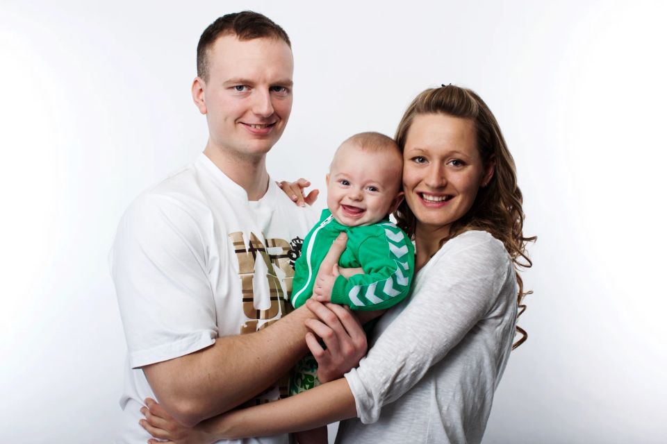 Sort-hvid familiefoto af mor, far og en glad baby, der smilende kigger mod kameraet i blødt studie-lys.