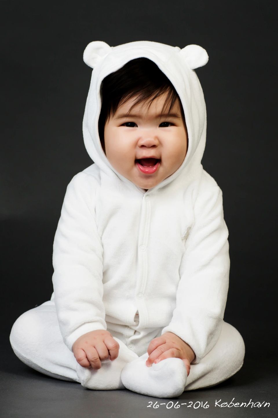 Glad baby i hvid bjørnedragt sidder foran mørk baggrund. Et dejligt babyfotografi med hjertevarm stemning.