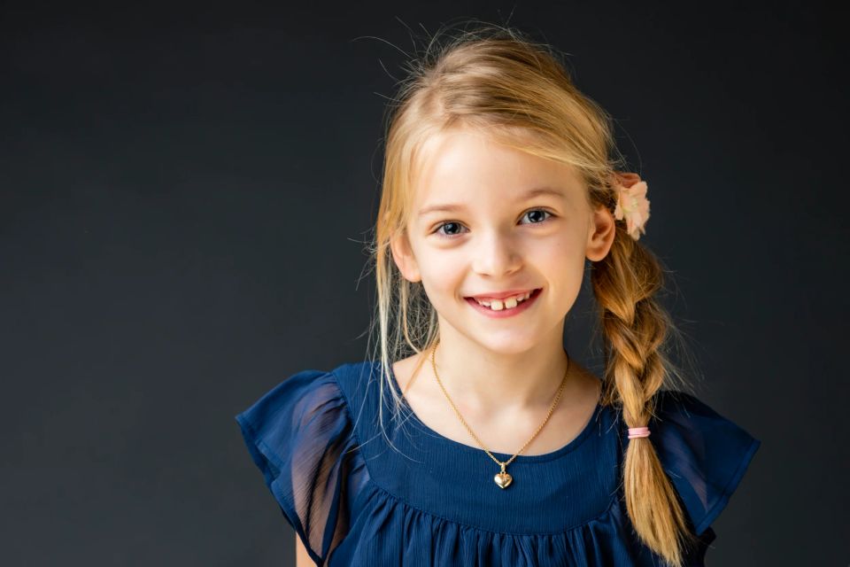 Glad portrætfoto af ung pige med lyst hår og fletning, smilende mod beskueren. Naturlig og varm stemning.