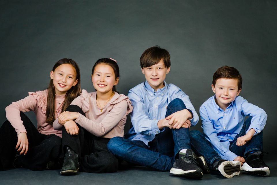 Børnefoto: Fire glade børn i lyse farver, siddende tæt sammen foran en mørk grå baggrund.