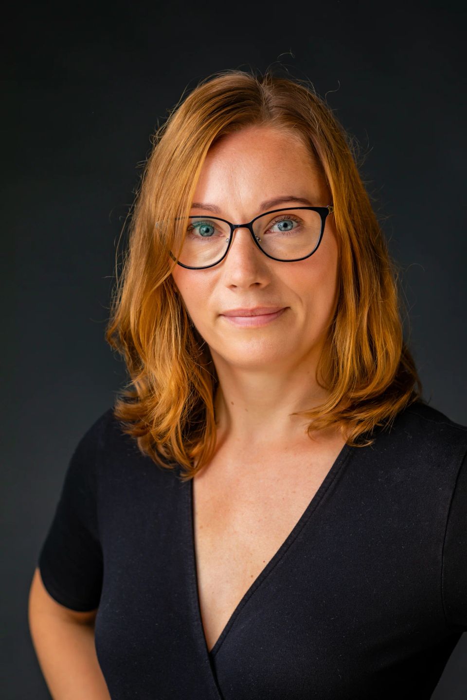 Professionelt portrætfoto af rødhåret kvinde med briller, der ser direkte ind i kameraet med et venligt udtryk.