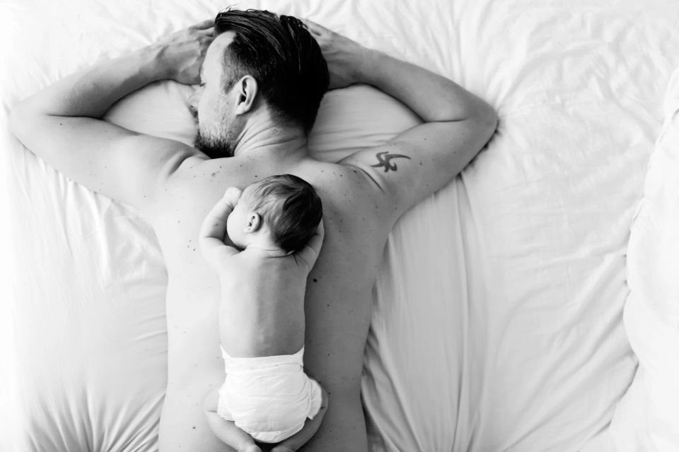 Far og baby sover trygt side om side i sort-hvidt babyfotografi, set oppefra. En rolig stemning.