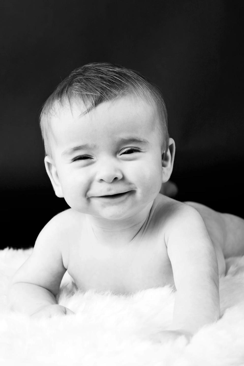 Babyfotografering af glad baby på hvidt tæppe. Babyen smiler bredt med lukkede øjne mod en mørk baggrund.