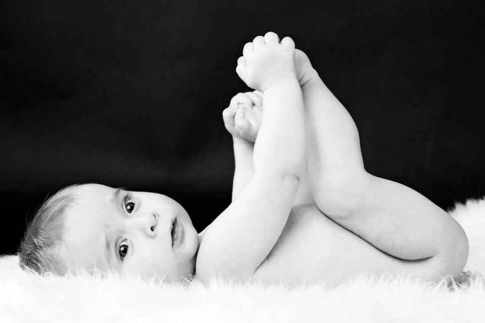 Sort-hvidt babyfoto: Nysgerrig baby ligger på ryggen på et blødt lammeskind med fødderne oppe mod en mørk baggrund.