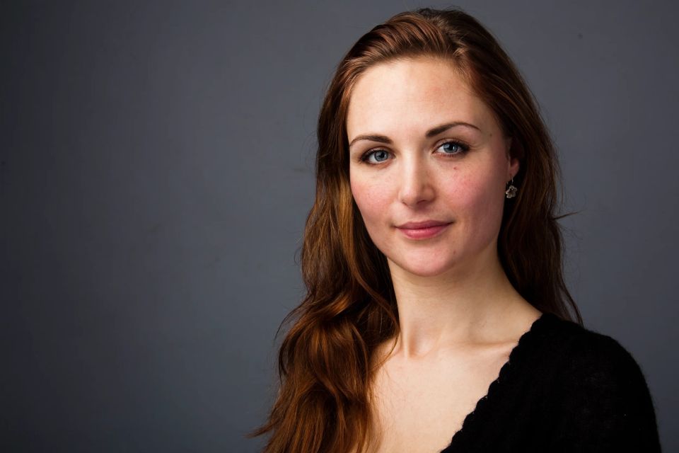 Portræt af en ung kvinde med langt brunt hår og blide, blå øjne, fotograferet i et naturligt, dæmpet lys mod en mørk baggr...