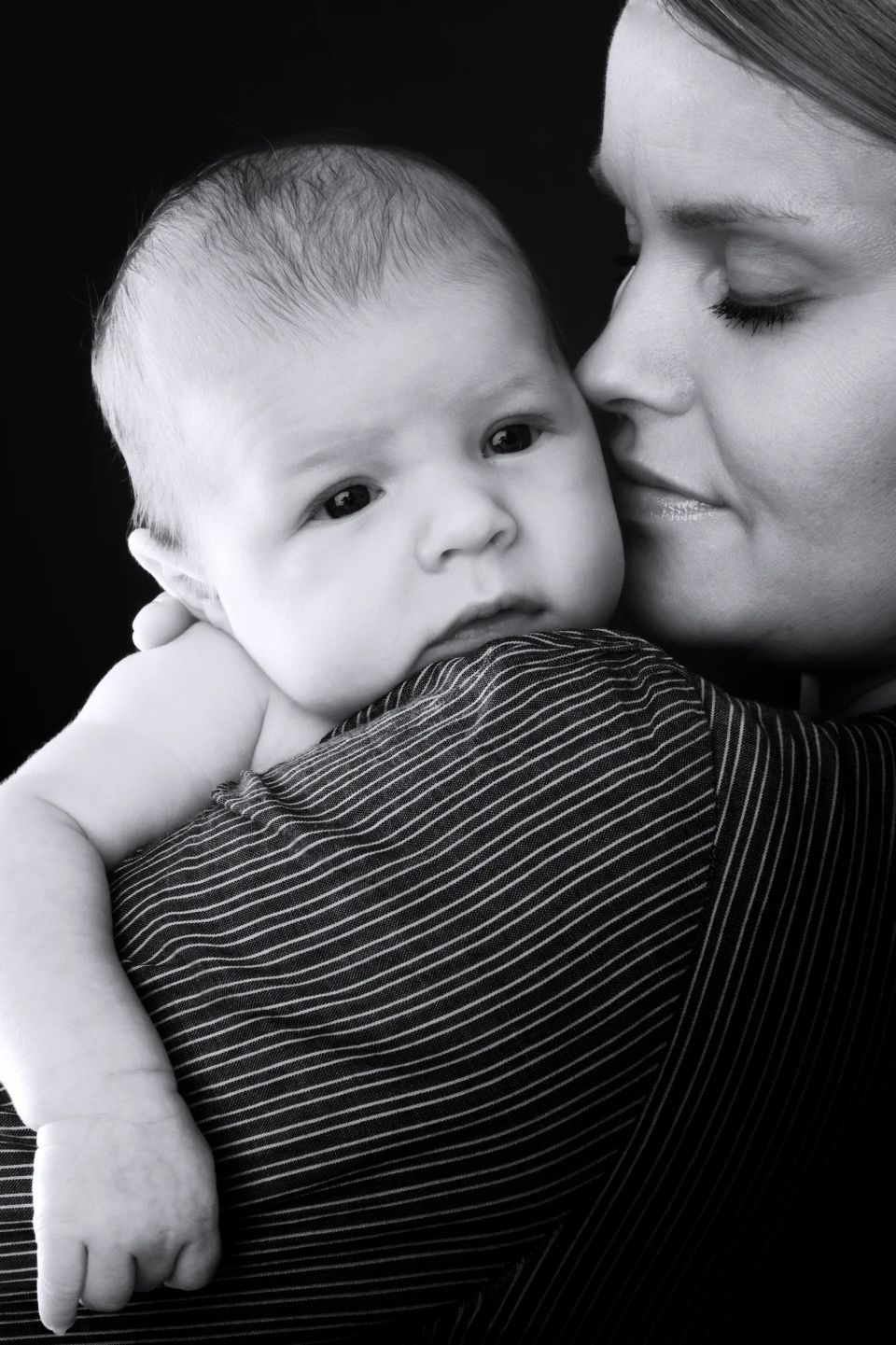 Sort-hvid babyfotografering: mor holder sin nyfødte tæt, kysser blidt med lukkede øjne i naturligt lys.