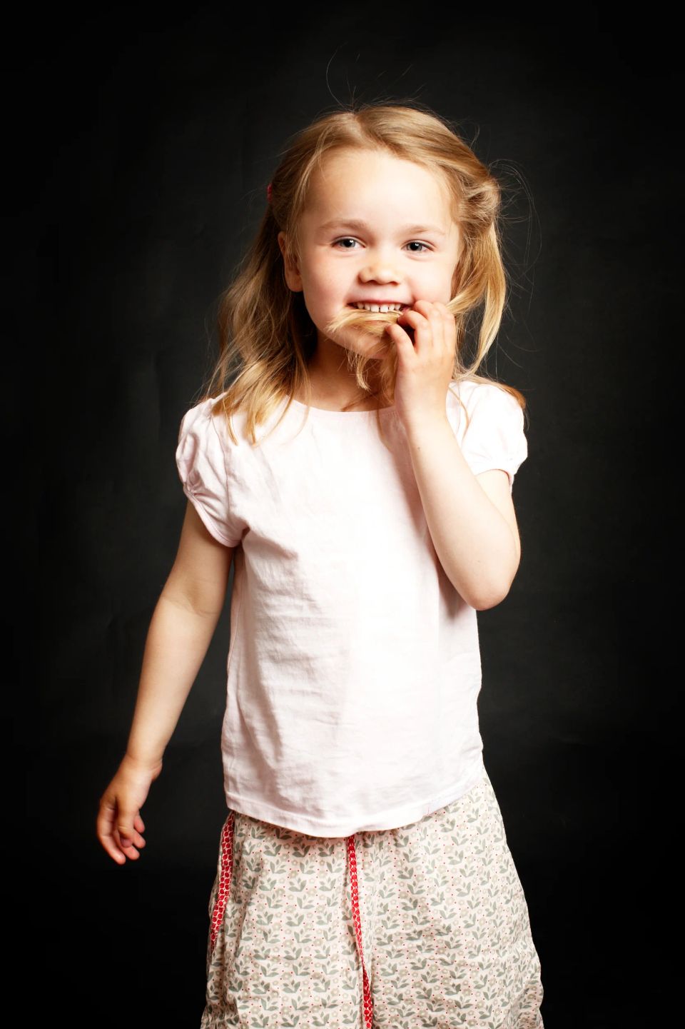 Børn: Et legende barn i lyserød bluse og mønstrede shorts smiler, med ansigtet omkranset af lyst hår mod mørk baggrund.