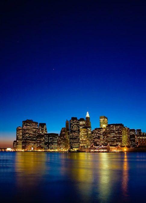 Oplyst Manhattan skyline set over vand ved daggry eller aften i mørkeblå toner.
