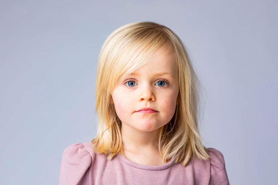 Børnefotografering af pige med blondt hår og intense blå øjne, neutral baggrund.