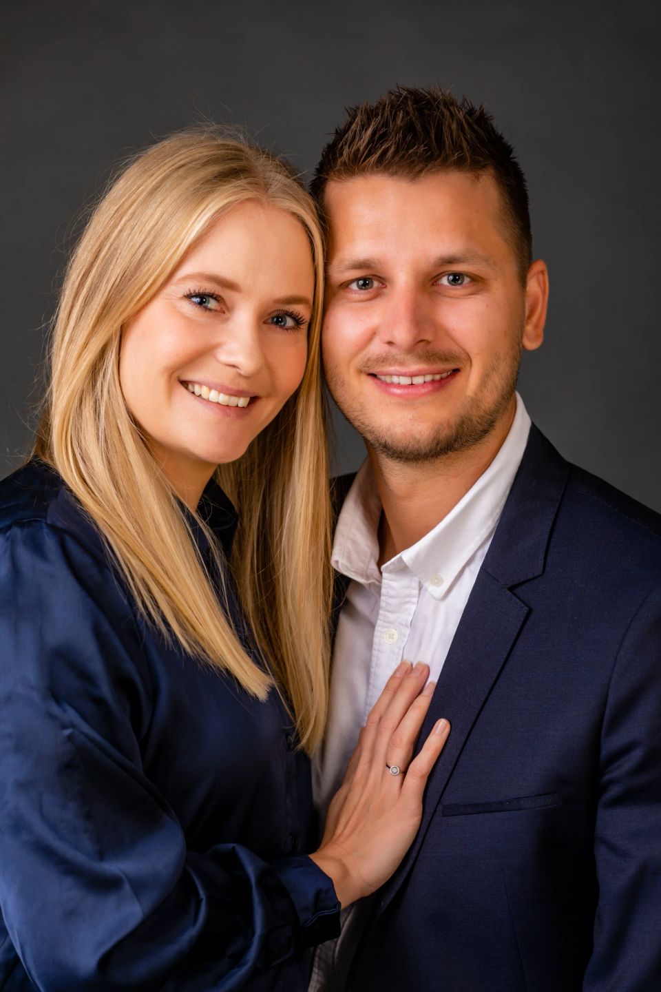 Gladt ungt par med blond kvinde i mørkeblå kjole, der smilende læner sig ind mod mand i jakkesæt. Et kærligt portrætfoto.