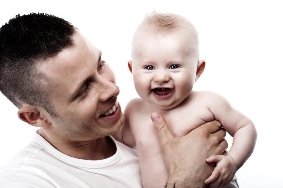 Baby fotografering hvor en far smiler varmt til sin glade baby, der griner og kigger mod kameraet i skarpt lys.