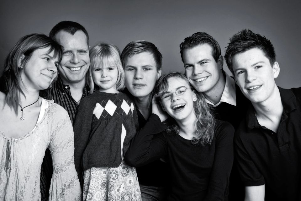 Sort-hvidt familiefoto af voksne og fire børn, der smilende poserer tæt sammen i blødt lys. Skaber en autentisk stemning.