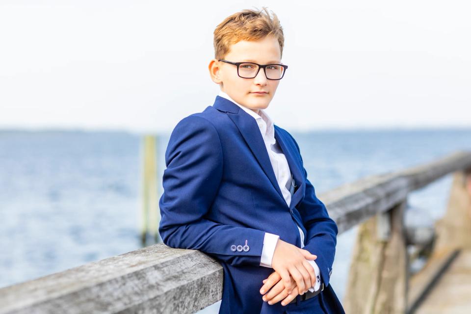 Portrætfotografering af konfirmand i blåt jakkesæt med briller, stående ved et trægelænder ud til vandet i dagslys.
