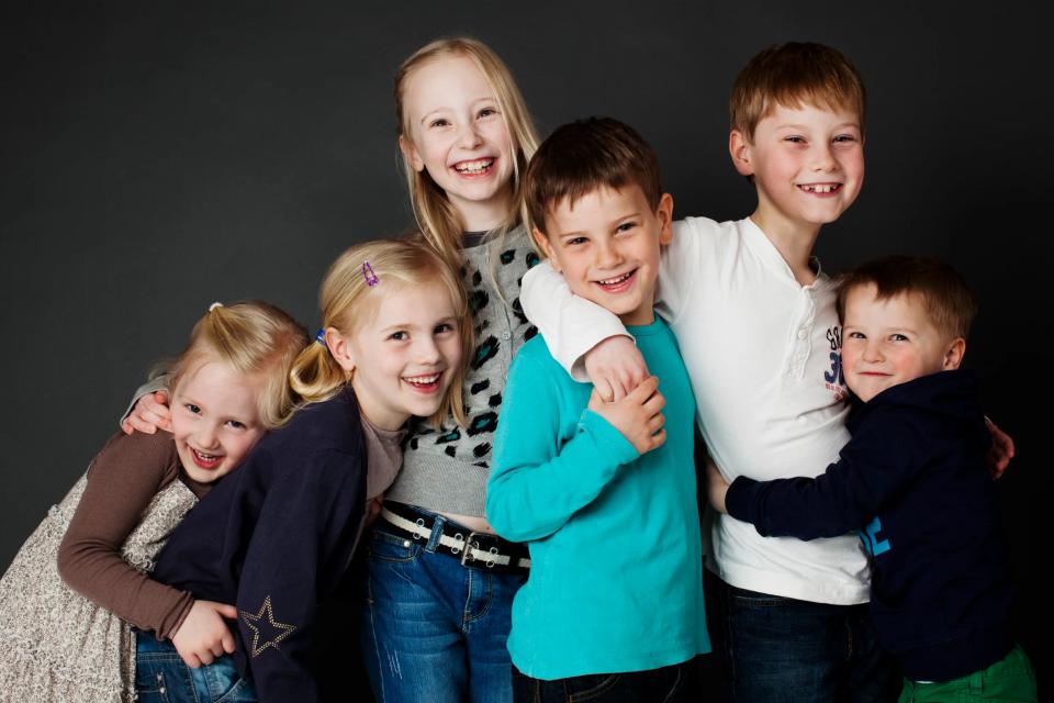 Familiefoto af seks glade, smilende børn tæt sammen mod en mørk baggrund, der fremhæver deres ansigter.
