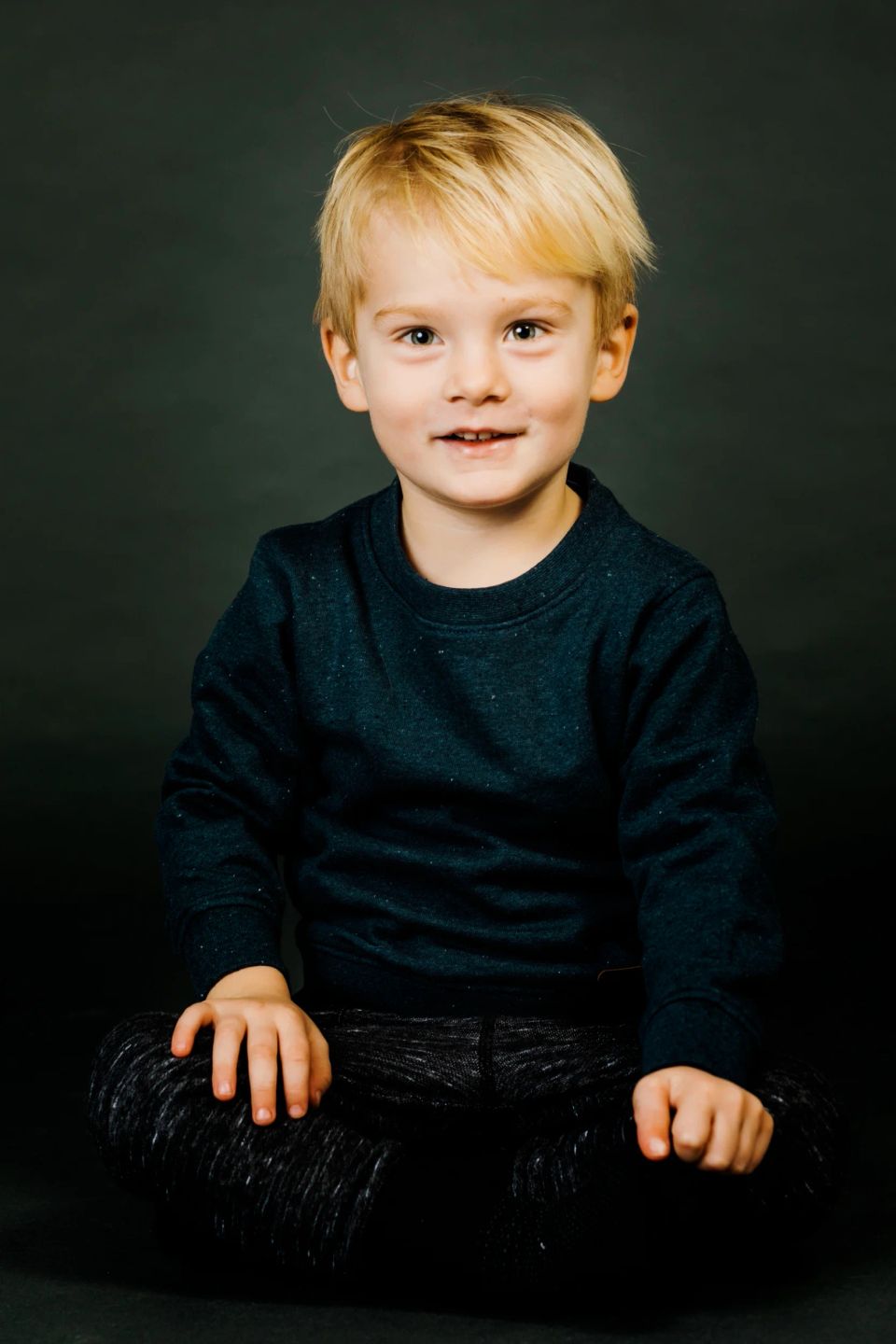 Børnefoto af blond dreng i mørkeblå trøje, der smiler charmerende til kameraet med et glimt i øjet.