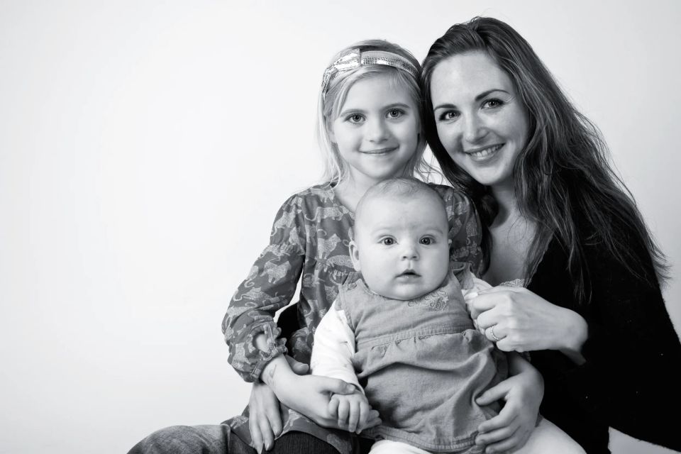 Sort-hvid familiefoto af smilende mor, der holder sin ældste datter og baby. Blødt naturligt lys.