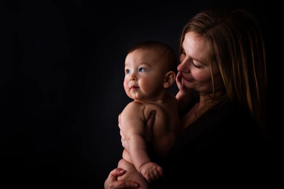 Nært babyfoto af smilende mor, der holder sin baby. Baby kigger nysgerrigt mod oplyst dybde i blødt sidelys.