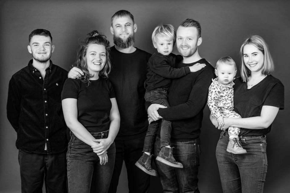 Familiefoto i sort-hvid af tre mænd, to kvinder, en lille dreng og en baby på armen, stående side om side.