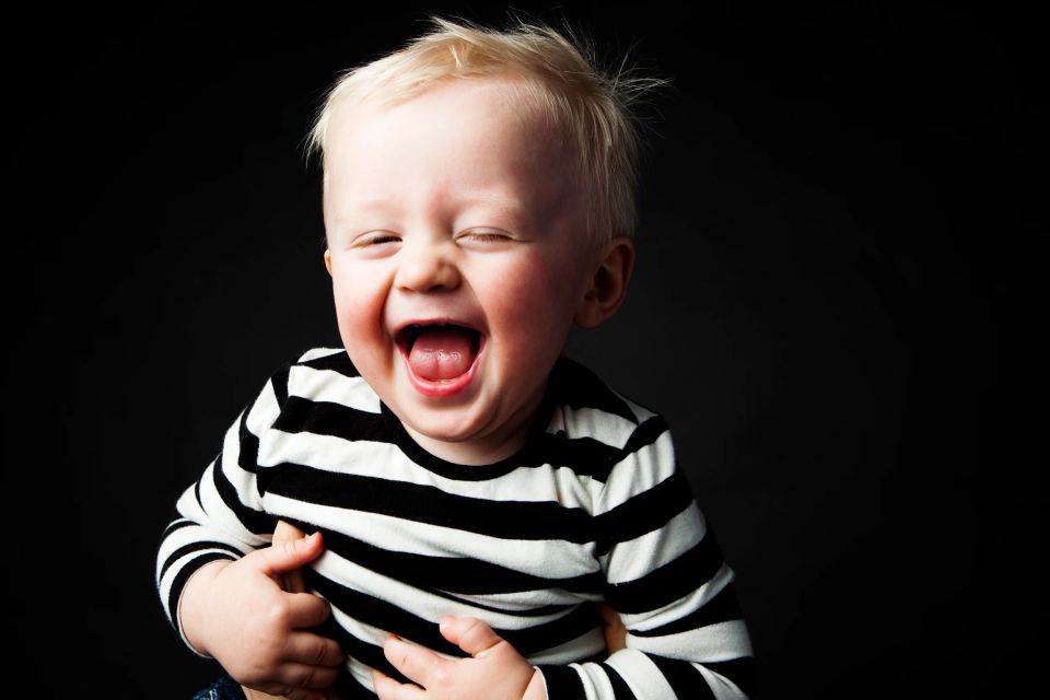 Glad babyfotografering: lysthåret baby i stribet bluse griner hjerteligt med lukkede øjne på mørk baggrund.