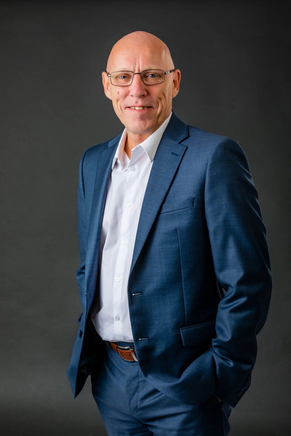 Glad mand i blåt jakkesæt og hvid skjorte med hænder i lommerne. Professionelt studieportræt i dagslys.