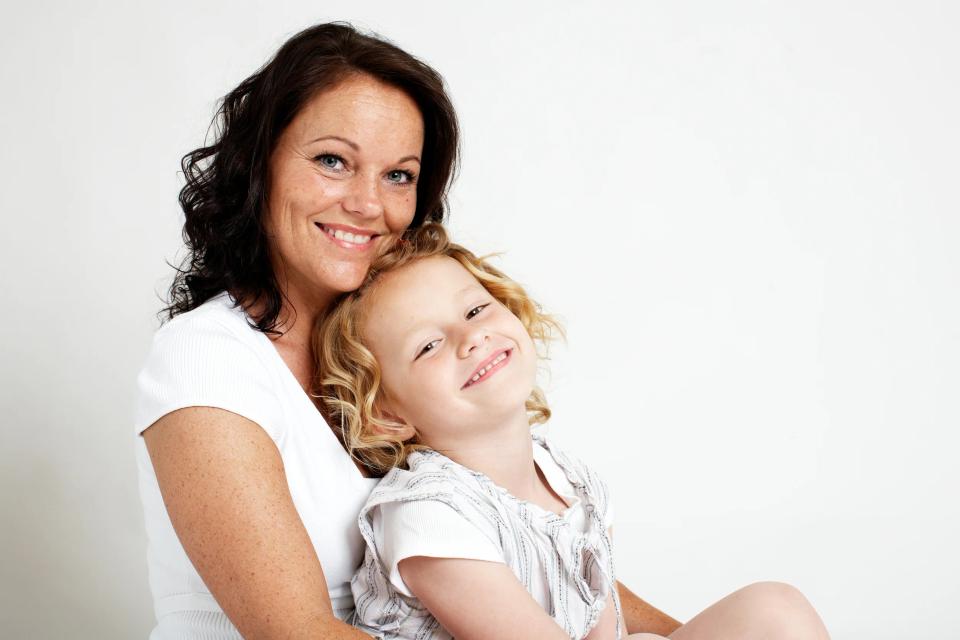 Familieportræt af smilende mor med mørkt hår, der krammer sin lille datter med krøllet lyst hår. Hjertevarm stemning.