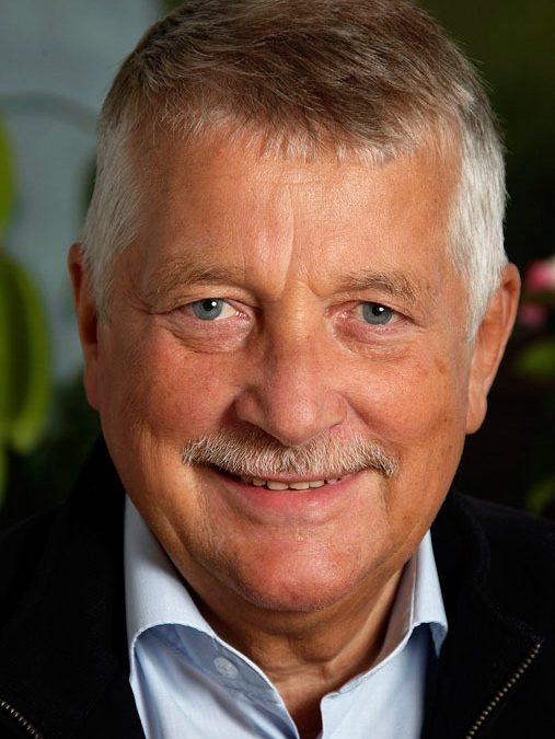 Autentisk portrætfoto af ældre smilende mand med gråt hår og overskæg i blød belysning. Blå skjorte, sort jakke.