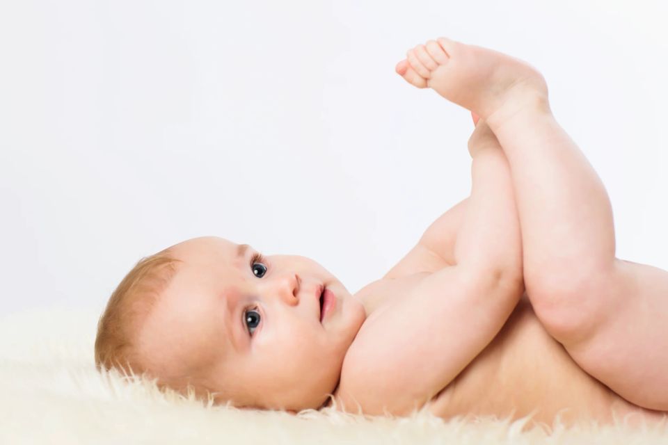 Legesyg baby med blå øjne liggende på ryg med løftede ben på et blødt, lyst tæppe. Babyfotografering i Virum.