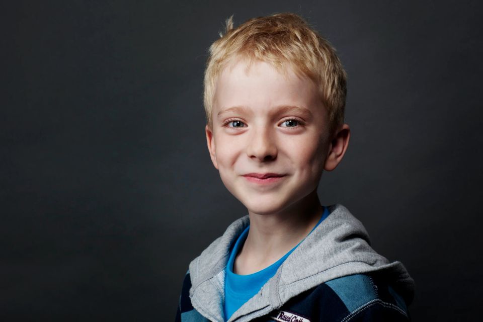 Portrætfoto af glad lyshåret dreng i hættetrøje mod en mørk baggrund, giver en varm og venlig stemning.